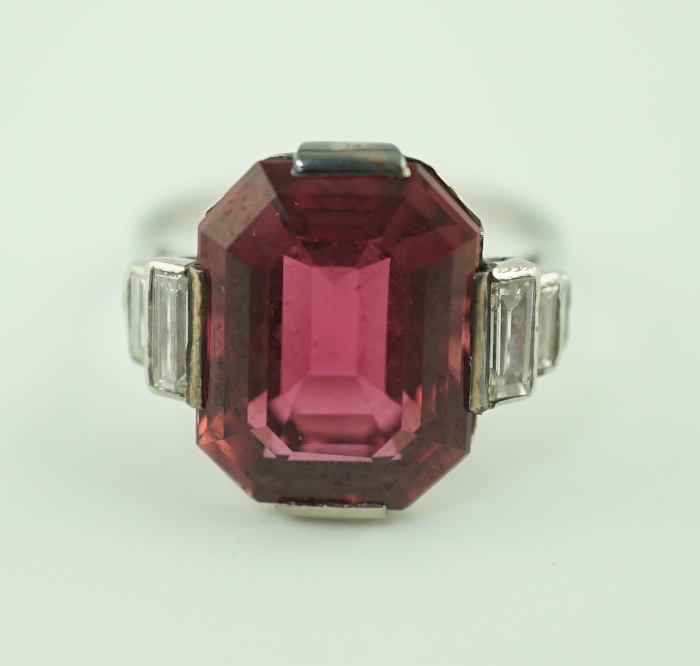 An Art Deco platinum and single stone emerald cut deep pink tourmaline set dress ring, with graduated six stone baguette cut diamond set shoulders